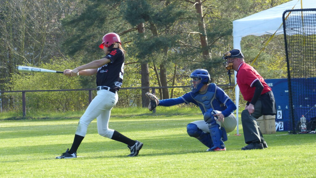 Baseballspieler in schwarzem trikot (rostock bucaneros) und rotem helm beim schlag, während der plate umpire in rot, hinter dem catcher in blau steht und beobachtet.