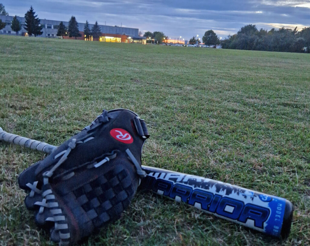Ein baseballhandschuh und ein baseballschläger liegen auf dem gras im vordergrund, während im hintergrund der dämmerungshimmel über einem ruhigen park mit beleuchteten gebäuden zu sehen ist.